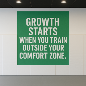 Motivational BJJ sign reading 'Growth starts when you train outside your comfort zone' displayed in a martial arts academy with white wall mats, gray floors, and black ceiling lighting.